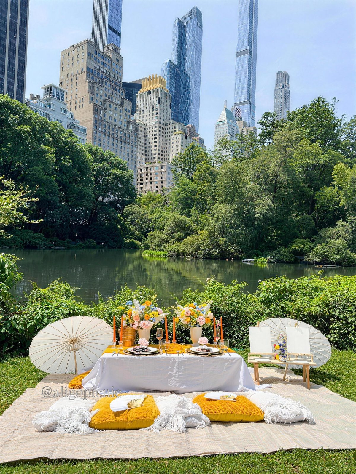 A white low table with a marigold runner, wildflowers and paper parasols beside the lake in Central Park
