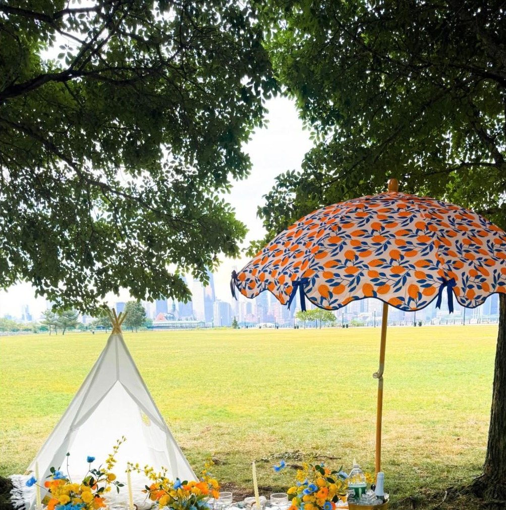 A white teepee and an orange print parasol on a wide lawn, with the city skyline beyond the trees
