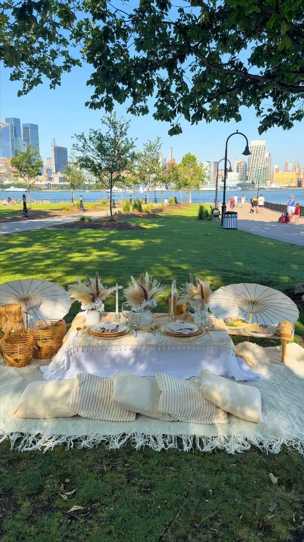 A cream picnic setup under the trees on the Hudson waterfront, with the Manhattan skyline across the river
